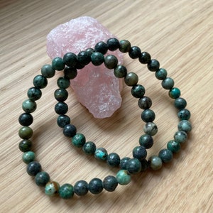 Può includere: Un braccialetto di perline di turchese con venature verde scuro e nero. Il braccialetto è fatto di perline rotonde e si trova su una superficie di legno.