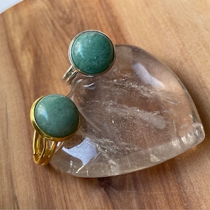 Puede incluir: Dos anillos de oro con engastes de piedras preciosas verdes. Los anillos están sobre una superficie de cristal transparente en forma de corazón.