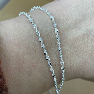 May include: Two silver chain bracelets with a delicate, twisted design. The bracelets are layered on a person's wrist.