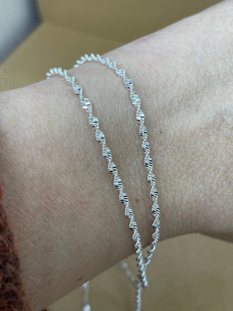 May include: Two silver chain bracelets with a delicate, twisted design. The bracelets are layered on a person's wrist.
