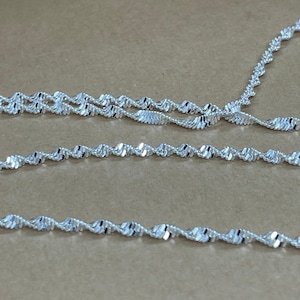May include: A silver chain necklace with a twisted or rope design. The chain is layered on a brown surface.
