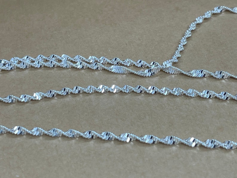 May include: A silver chain necklace with a twisted or rope design. The chain is layered on a brown surface.