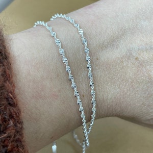 May include: Two silver chains with a twisted design, worn on a wrist.