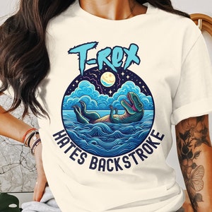 Puede incluir: Una camiseta blanca con un gráfico azul y verde de un dinosaurio nadando en el océano. El gráfico incluye el texto "T-Rex Hates Backstroke".