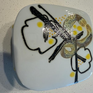 May include: A square white ceramic box with rounded corners featuring a hand-painted design of black outlined flowers, yellow dots, and textured brown and green circular accents on the lid, showcasing a glossy finish and a clean, smooth surface.