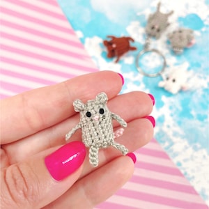 May include: A small gray crocheted mouse with pink ears and a pink nose. The mouse is being held in a hand.