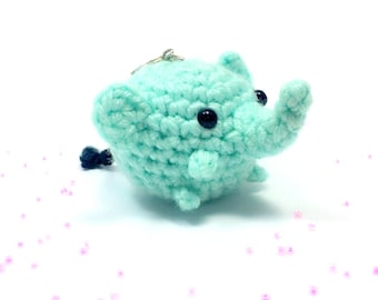 May include: A small, turquoise crocheted elephant keychain with black eyes and a silver keyring. The elephant has a trunk, ears, and a small tail. The background is white with a few pink specks.