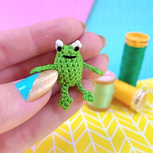 May include: A small green crocheted frog with white eyes. The frog is being held in a hand with blue and gold nail polish.