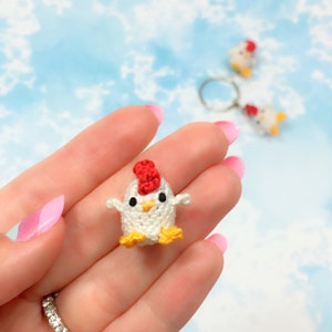 May include: A small white crocheted chicken with a red comb and yellow feet. The chicken is being held in a hand.