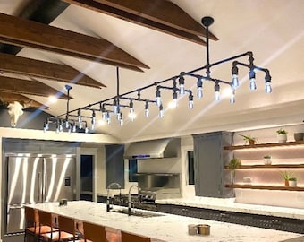 May include: A modern kitchen with a long, industrial-style light fixture with exposed bulbs hanging over a white marble kitchen island with bar stools. The kitchen has stainless steel appliances and gray cabinets.