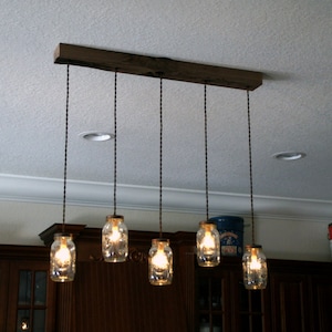 May include: A rustic, wooden beam ceiling light fixture with five mason jar pendant lights. The jars are clear, and the bulbs inside are lit, casting a warm glow. The fixture is mounted on a white ceiling.