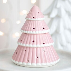 Può includere: Un portacandele a forma di albero di Natale in ceramica rosa. L'albero ha tre livelli con ritagli a forma di stella e finiture bianche. L'albero è su una superficie bianca, con uno sfondo sfocato.