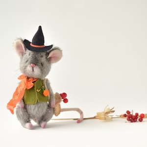 May include: A gray plush mouse wearing a black witch hat with an orange band, a green vest, and an orange scarf. The mouse is holding a small brown object and is sitting on a white surface. There are dried flowers and red berries in the background.