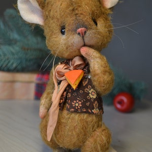 Op de afbeelding: Een bruine en witte pluche muis die een bruin vest draagt met bloemenprint en een perzikkleurig lint met een klein oranje en geel stukje kaas. De muis houdt zijn pootje bij zijn mond en heeft een roze neus.