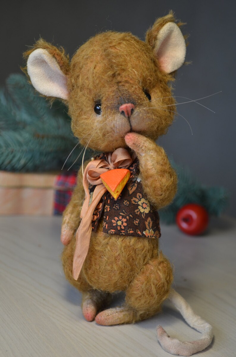 Op de afbeelding: Een bruine en witte pluche muis die een bruin vest draagt met bloemenprint en een perzikkleurig lint met een klein oranje en geel stukje kaas. De muis houdt zijn pootje bij zijn mond en heeft een roze neus.
