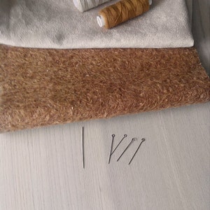 Op de afbeelding: Twee spoelen garen, een witte en een bruine, liggen op een beige stof. Een bruine stof ligt onder de beige stof. Vier spelden liggen op een licht houten oppervlak.