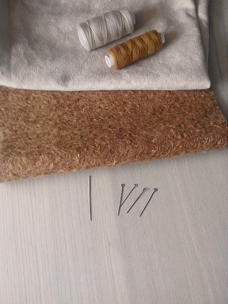 Op de afbeelding: Twee spoelen garen, een witte en een bruine, liggen op een beige stof. Een bruine stof ligt onder de beige stof. Vier spelden liggen op een licht houten oppervlak.