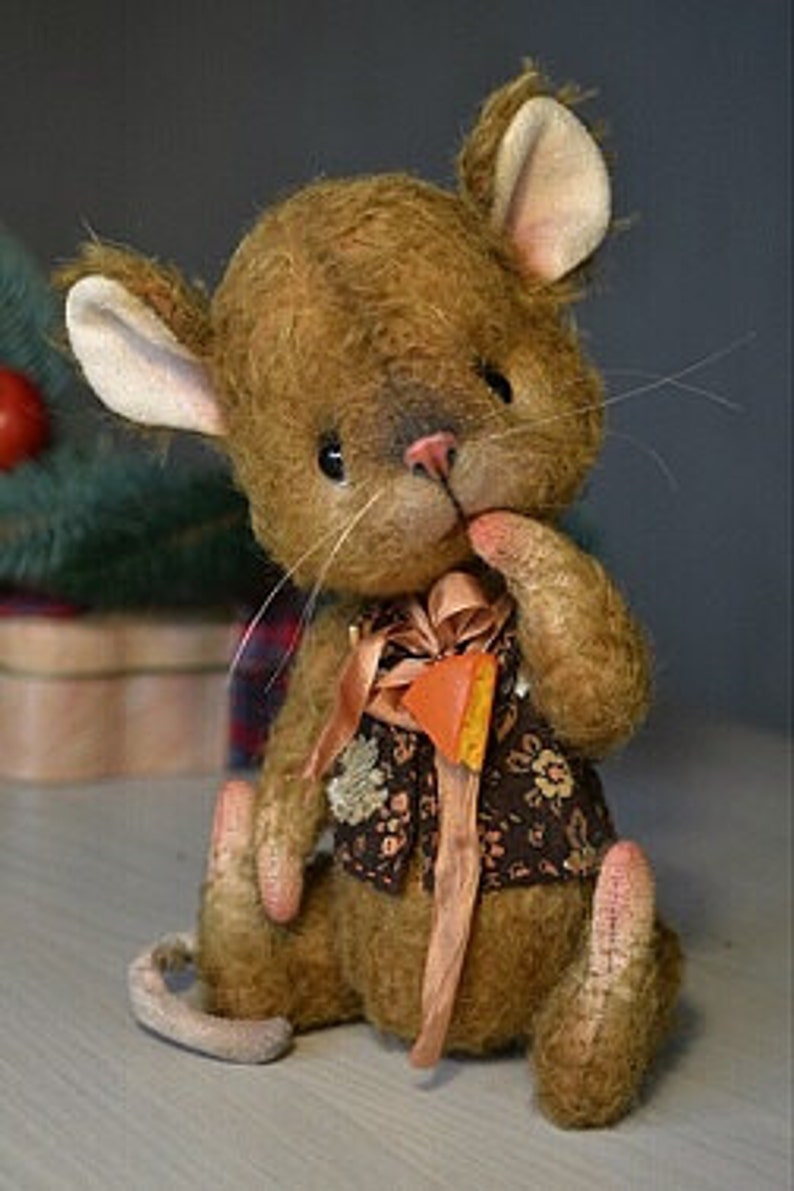 Op de afbeelding: Een bruine pluche muis die een bruin vest met bloemenprint en een bruin lint draagt. De muis heeft een lange staart en zit op een wit oppervlak.