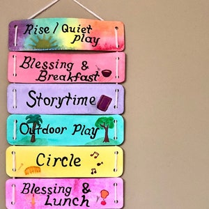 K&ouml;nnte beinhalten: Ein farbenfroher Wandbehang aus Holz mit bemalten Holzbl&ouml;cken. Jeder Block hat eine andere Farbe und zeigt eine andere Aktivit&auml;t, wie "Aufstehen / Ruhiges Spiel", "Segen und Fr&uuml;hst&uuml;ck", "Geschichtenzeit", "Spiel im Freien", "Kreis", "Segen und Mittagessen", "T&auml;gliche Aktivit&auml;t", "Spielen", "Nachmittagstee und Snack", "Ruhe", "Hauspflege", "Segen und Abendessen", "Familienzeit", "Bad", und "Gute Nacht".