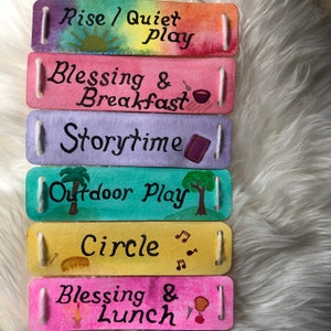 K&ouml;nnte beinhalten: Sechs bunte Filzstreifen mit gemalten Bildern und Text. Die Streifen lauten "Rise/Quiet Play", "Blessing & Breakfast", "Storytime", "Outdoor Play", "Circle" und "Blessing & Lunch".