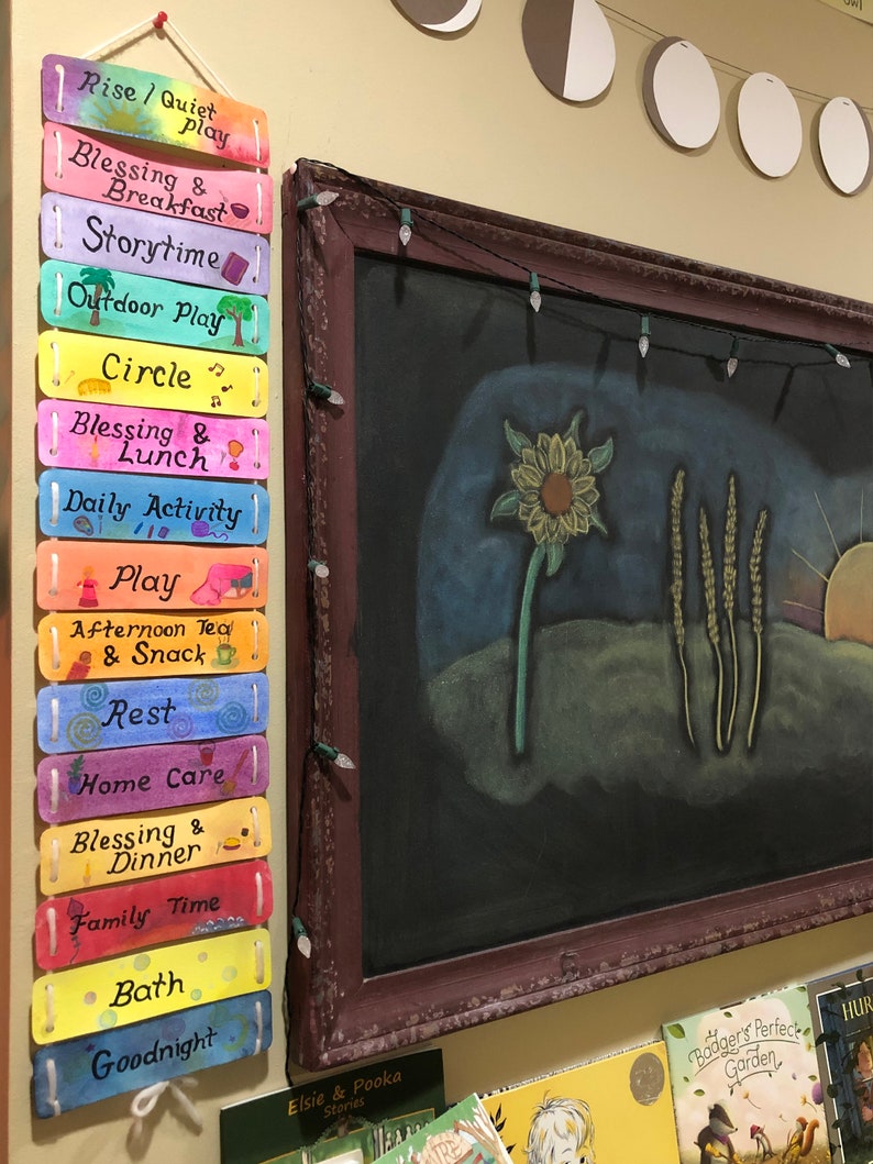 K&ouml;nnte beinhalten: Eine bunte Holztafel mit einer Schnur oben, die an einer Wand h&auml;ngt. Die Tafel hat 14 rechteckige Abschnitte, jeder in einer anderen Farbe und mit einem handschriftlichen Satz. Die S&auml;tze lauten "Rise/Quiet Play", "Blessing & Breakfast", "Storytime", "Outdoor Play", "Circle", "Blessing & Lunch", "Daily Activity", "Play", "Afternoon Tea & Snack", "Rest", "Home Care", "Blessing & Dinner", "Family Time" und "Goodnight".