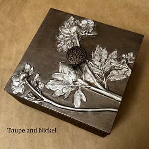 May include: A square taupe box featuring a raised nickel-colored floral relief design on the lid. The lid includes a small, circular, textured dark knob. The text "Taupe and Nickel" is printed in the bottom left corner of the image.