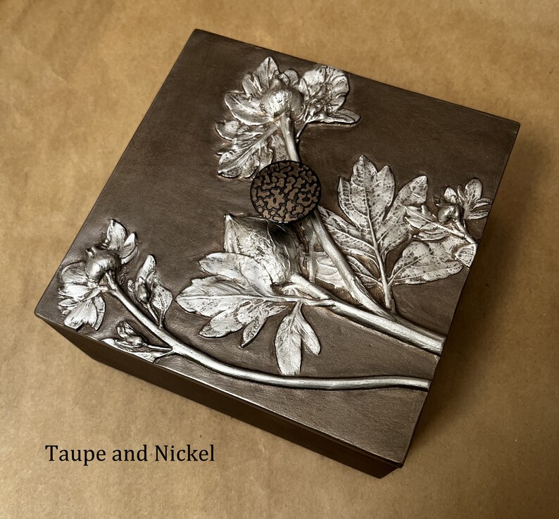 May include: A square taupe box featuring a raised nickel-colored floral relief design on the lid. The lid includes a small, circular, textured dark knob. The text "Taupe and Nickel" is printed in the bottom left corner of the image.