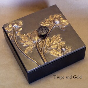 May include: A square box with a black base and a metallic taupe lid featuring a raised gold floral relief design. A textured dark circular knob is centered on the lid. The text Taupe and Gold is printed in the lower right corner of the image.