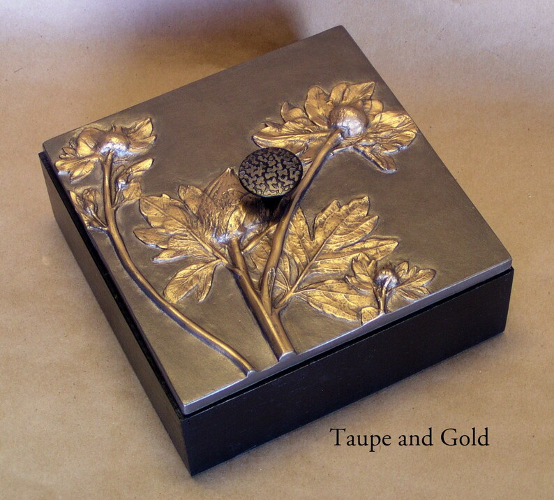 May include: A square box with a black base and a metallic taupe lid featuring a raised gold floral relief design. A textured dark circular knob is centered on the lid. The text Taupe and Gold is printed in the lower right corner of the image.