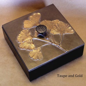 May include: A square box with a dark taupe lid featuring a raised gold ginkgo leaf design and a central dark textured knob. The sides of the box are black. The text Taupe and Gold is printed in the bottom right corner of the image.