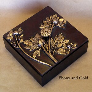 May include: A square dark brown wooden box featuring a raised metallic gold floral relief on the lid. The design depicts stems, leaves, and flower buds in a textured finish. The text Ebony and Gold is printed in the bottom right corner of the image.