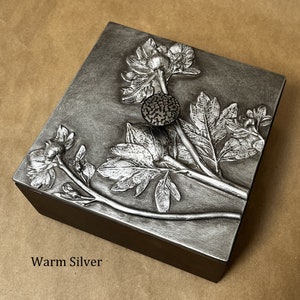 May include: A square box with a dark brown base and a metallic warm silver lid. The lid features a raised relief design of floral stems, leaves, and blossoms. A small, circular, textured dark button is centered on the lid. The text WARM SILVER is visible.