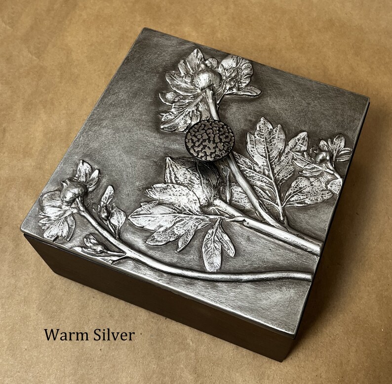 May include: A square box with a dark brown base and a metallic warm silver lid. The lid features a raised relief design of floral stems, leaves, and blossoms. A small, circular, textured dark button is centered on the lid. The text WARM SILVER is visible.
