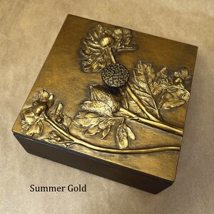 May include: A square wooden box with a metallic gold finish on the lid, featuring a raised relief of a floral stem with leaves and a textured circular knob. The text Summer Gold is printed in black on the lower left side of the box surface.