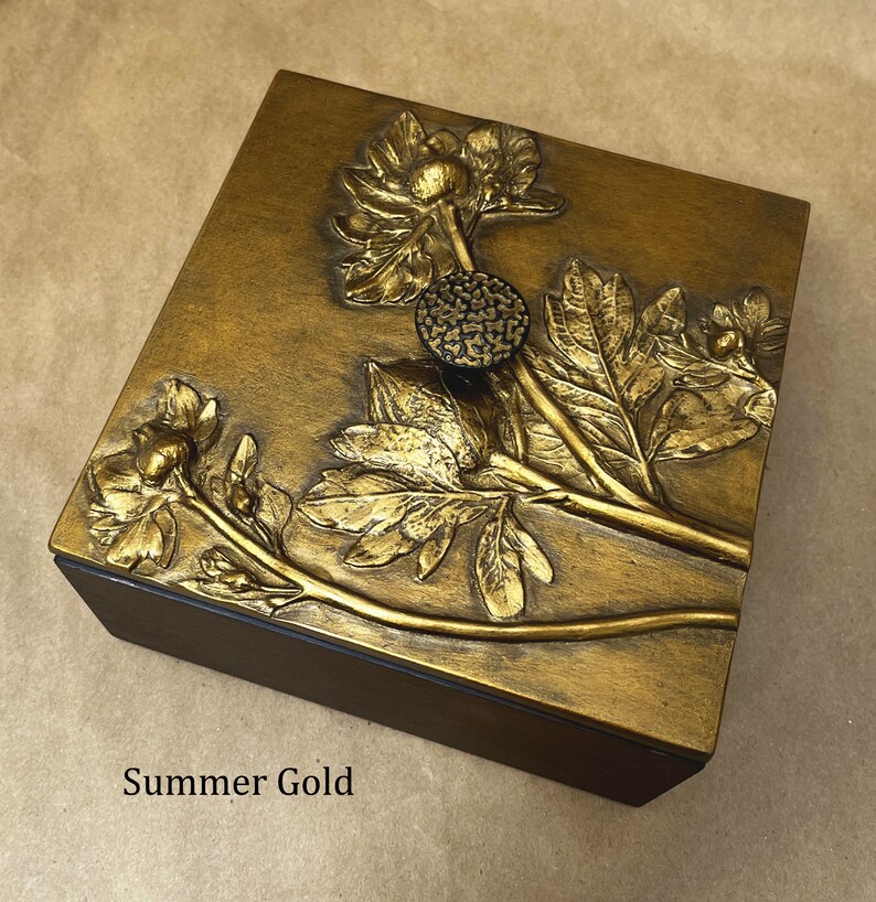 May include: A square wooden box with a metallic gold finish on the lid, featuring a raised relief of a floral stem with leaves and a textured circular knob. The text Summer Gold is printed in black on the lower left side of the box surface.