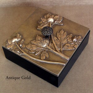 May include: A square box with a black base and an antique gold lid featuring a raised relief of floral stems, leaves, and blossoms. A small, round, textured black knob is centered on the lid. The text Antique Gold is printed below the box.