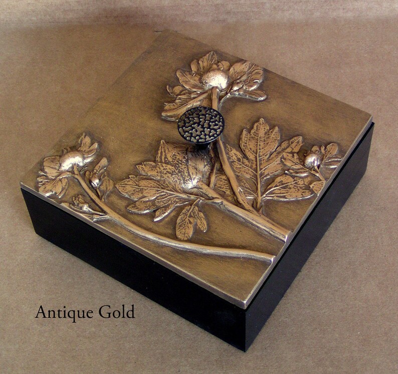 May include: A square box with a black base and an antique gold lid featuring a raised relief of floral stems, leaves, and blossoms. A small, round, textured black knob is centered on the lid. The text Antique Gold is printed below the box.
