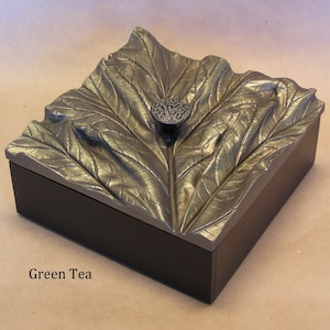 May include: A square wooden box with a gold leaf lid featuring a raised leaf design. The lid has a metal knob. The box is labeled "Green Tea".