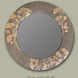 May include: A round wall mirror with a taupe frame featuring raised gold-colored ginkgo leaf branch accents. The mirror has a central circular reflective glass surface. The text "Taupe and Gold 30"" is printed below the mirror frame.