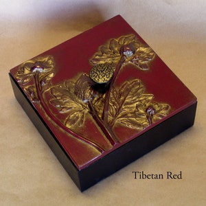 May include: A square box with a black base and a Tibetan Red lid featuring a raised gold floral relief design. A textured gold knob is centered on the lid. The text Tibetan Red is printed in the lower right corner of the image area.