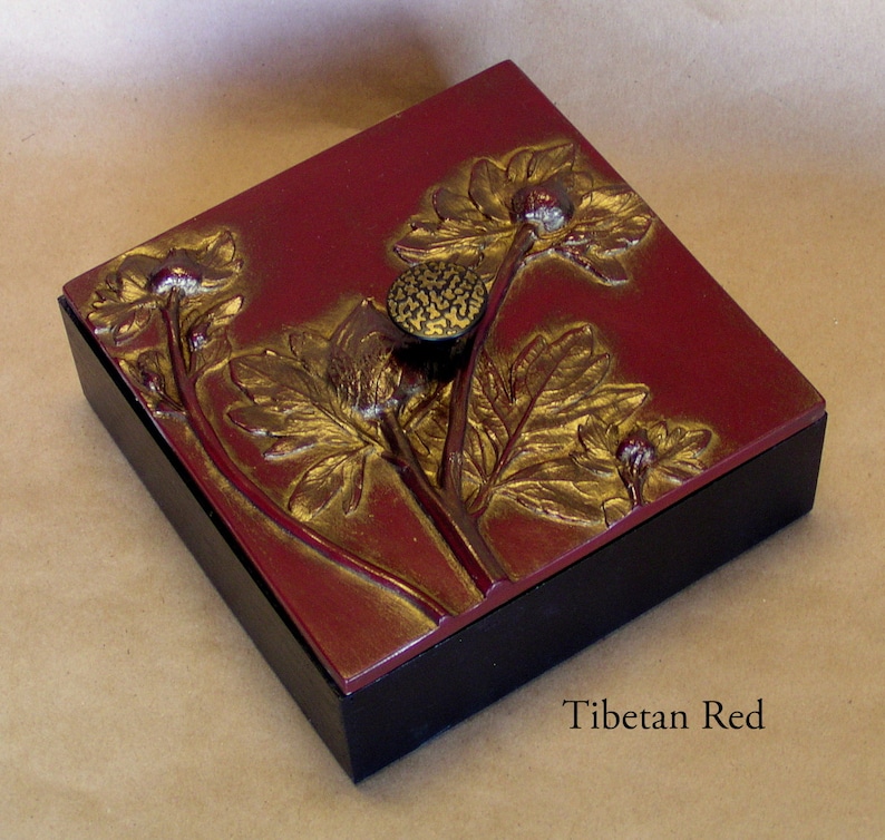 May include: A square box with a black base and a Tibetan Red lid featuring a raised gold floral relief design. A textured gold knob is centered on the lid. The text Tibetan Red is printed in the lower right corner of the image area.