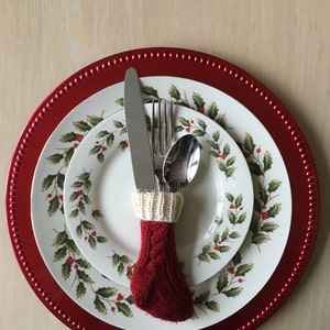 May include: A red and white knitted stocking holds a fork, knife, and spoon on a white plate with a holly pattern. The plate is on a red charger plate.