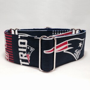 May include: A navy blue fabric pet collar featuring a repeating pattern of the New England Patriots logo in red, white, and blue, along with the word Patriots in white text. The collar includes silver-toned metal hardware for adjustment.