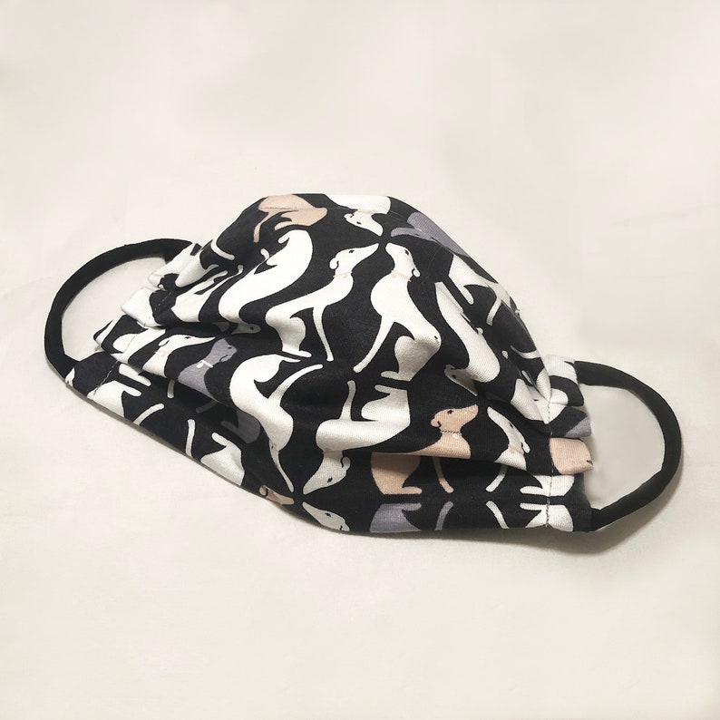 May include: A black face mask with a repeating pattern of white, gray, and tan dog silhouettes. The mask has black ear loops and is made of a pleated fabric.