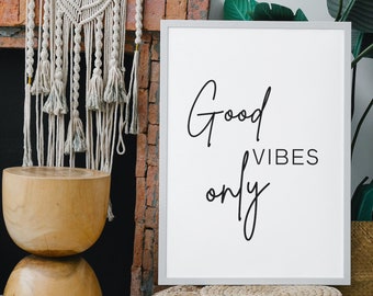 May include: White framed print with the text "Good Vibes Only" in black script font. The print is leaning against a brick fireplace with a macrame wall hanging and a wooden stool. A woven ottoman sits in front of the print.