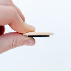 Pu&ograve; includere: Un magnete rotondo in legno con un retro magnetico nero. Il magnete &egrave; tenuto da una mano.