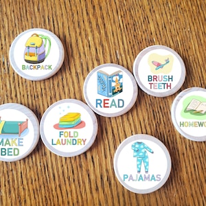 Puede incluir: Siete fichas redondas de madera con ilustraciones y texto coloridos. Las fichas presentan imágenes de una mochila, un cepillo de dientes, un libro, ropa doblada, una cama, pijamas y un libro abierto con un lápiz. El texto en las fichas dice "BACKPACK", "BRUSH TEETH", "READ", "FOLD LAUNDRY", "MAKE BED", "PAJAMAS", y "HOMEWORK".