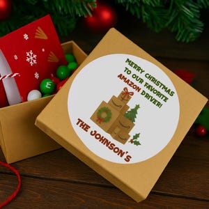 May include: A tan gift box with a white circular sticker that reads "MERRY CHRISTMAS TO OUR FAVORITE AMAZON DRIVER!" and "THE JOHNSON'S". The box contains a red gift box with a snowflake design and green and white candies.