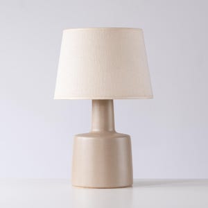 May include: A beige table lamp with a cylindrical base and a slender neck supporting a tapered lampshade. The lampshade is a light beige color, and the lamp has a minimalist design. The lamp is suitable for a bedroom or living room.