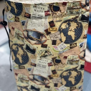 May include: A beige drawstring bag with a vintage travel theme print. The bag features a repeating pattern of maps, globes, and travel-related imagery. The text "ROUTE MAPS" is visible on the bag.